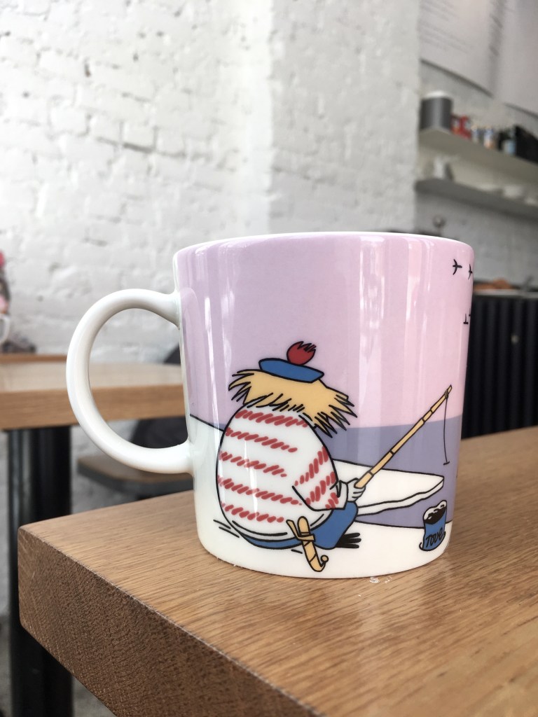 This colour photo shows a pink mug on a cafe table. The mug features the Moomin character Too-ticky.